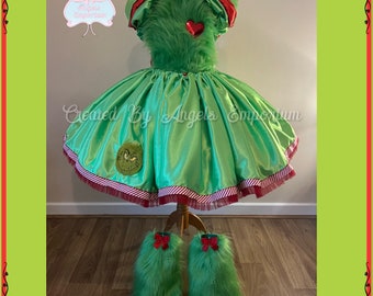 May include: Green satin dress with red trim and a Grinch face applique. The dress has a fuzzy green bodice and red heart accents. The dress is paired with green fuzzy leg warmers.