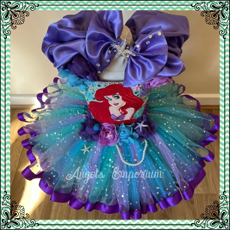 Puede incluir: Un vestido de tut&uacute; de tul turquesa y morado con un acabado brillante. El vestido presenta una aplicaci&oacute;n de fieltro de Ariel de La Sirenita, un lazo de sat&eacute;n morado y un collar de perlas. El vestido est&aacute; etiquetado como "Angels Emporium".