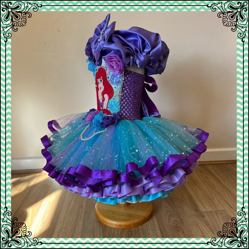 Puede incluir: Un tut&uacute; de tul azul y morado con un corpi&ntilde;o de sat&eacute;n morado y un lazo de sat&eacute;n morado. El corpi&ntilde;o presenta una imagen bordada en rojo y blanco de Ariel de La Sirenita. El tut&uacute; tiene capas de tul y cinta de sat&eacute;n.