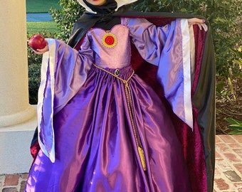 May include: A purple and black costume with a gold crown and a red apple. The costume features a long, flowing purple skirt, a bodice with a red jewel, and long sleeves. The cape is black with a red lining. Text on the bottom reads "Created By Angels Emporium."