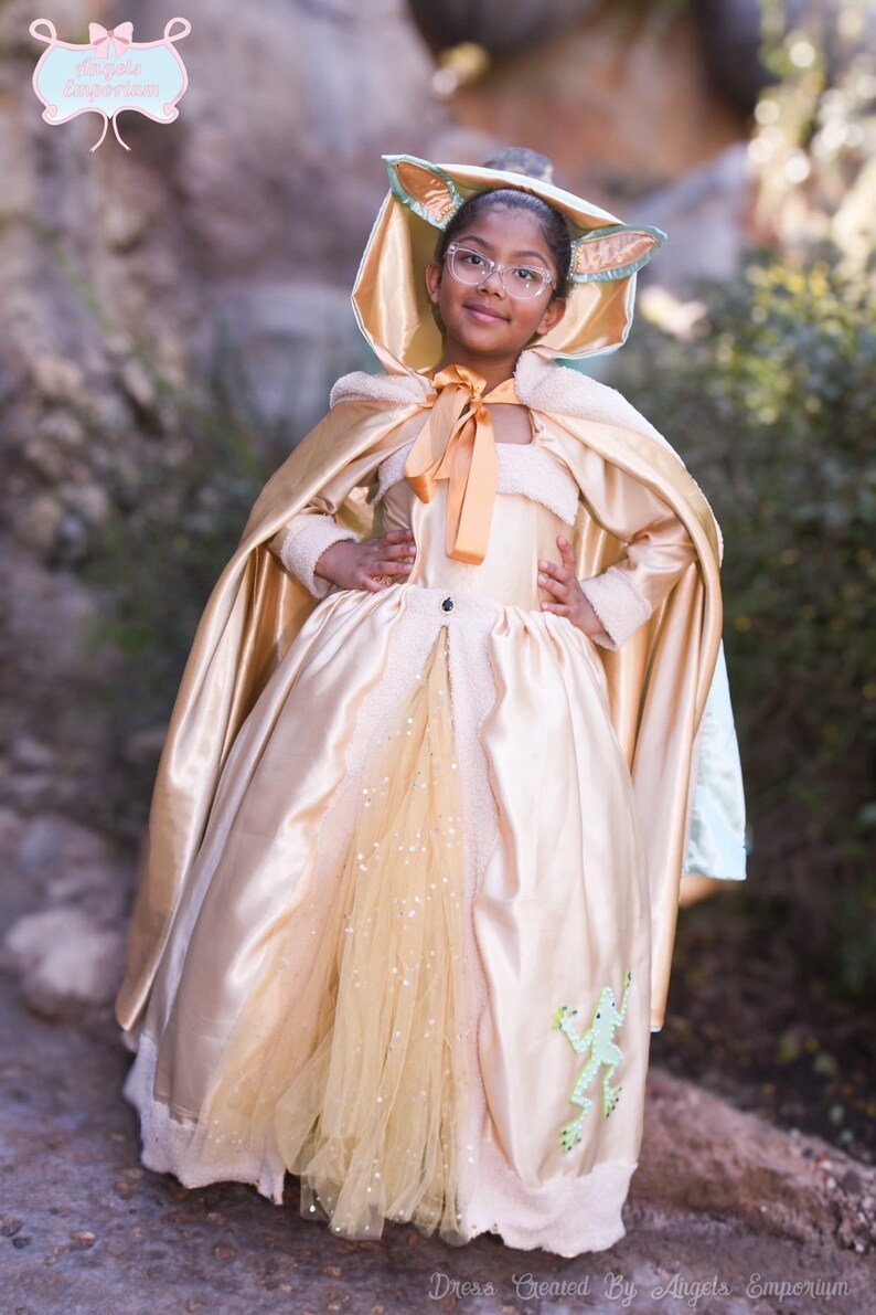 Puede incluir: Una ni&ntilde;a peque&ntilde;a con un disfraz de princesa dorado y verde con una capa a juego. El vestido tiene una falda de tul y una aplicaci&oacute;n de rana. La capa tiene una capucha con orejas.