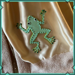 Puede incluir: Una aplicaci&oacute;n de rana verde con adornos brillantes verdes y blancos sobre un fondo de tela dorada clara.