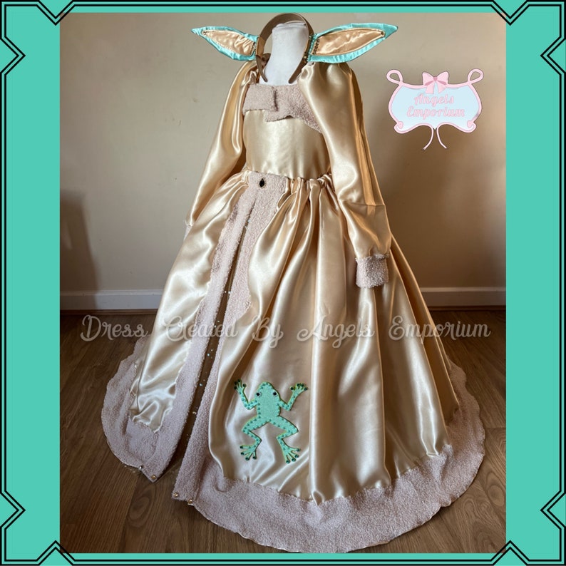 Puede incluir: Un vestido de sat&eacute;n dorado con ribete de piel marr&oacute;n y una aplicaci&oacute;n de rana verde. El vestido tiene una diadema a juego con orejas verdes. El vestido est&aacute; etiquetado como "Dress Created By Angels Emporium".