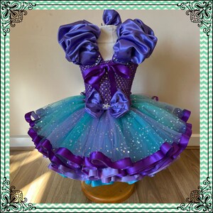 Puede incluir: Un vestido de tul de color morado y azul con un corpi&ntilde;o de sat&eacute;n y mangas abullonadas. El vestido est&aacute; decorado con una cinta morada y una estrella de mar.