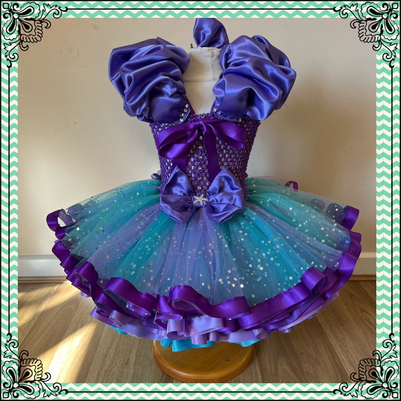 Puede incluir: Un vestido de tul de color morado y azul con un corpi&ntilde;o de sat&eacute;n y mangas abullonadas. El vestido est&aacute; decorado con una cinta morada y una estrella de mar.