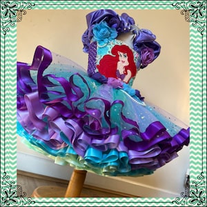 Puede incluir: Una falda de tul de varias capas con volantes de cinta de sat&eacute;n morado, azul y verde. La falda tiene un corpi&ntilde;o de lentejuelas con una imagen bordada de Ariel de La Sirenita.
