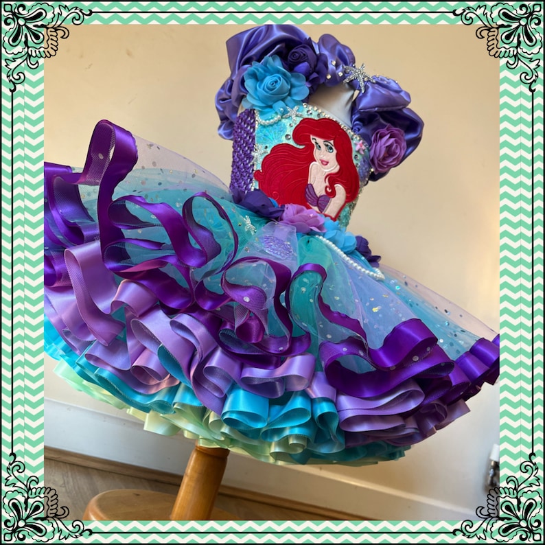 Puede incluir: Una falda de tul de varias capas con volantes de cinta de sat&eacute;n morado, azul y verde. La falda tiene un corpi&ntilde;o de lentejuelas con una imagen bordada de Ariel de La Sirenita.