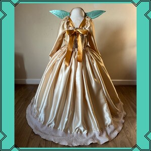 Puede incluir: Un vestido de sat&eacute;n dorado con un gran lazo y un dobladillo con volantes. El vestido tiene mangas largas y un escote en V. El vestido est&aacute; en un maniqu&iacute; con orejas verdes.