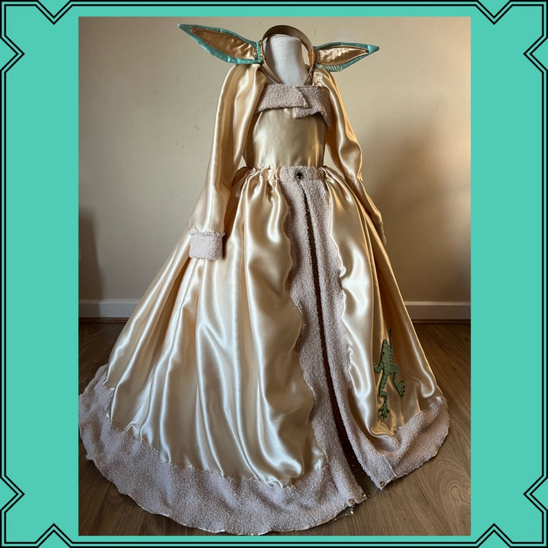 Puede incluir: Un vestido largo de sat&eacute;n dorado fluido con ribete de piel marr&oacute;n. El vestido tiene una falda larga y fluida y un corpi&ntilde;o ajustado. El vestido est&aacute; decorado con una aplicaci&oacute;n de rana verde en la falda.