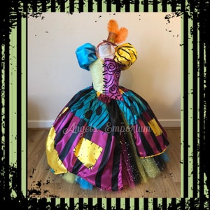 May include: A multi-coloured patchwork dress with a black and white striped bodice, yellow and black striped sleeves, and a blue tulle skirt. The dress is decorated with black stitching and has a black bow at the waist. The dress is worn by a mannequin with a black and orange hat with feathers.