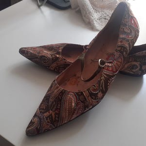May include: A pair of pointed-toe flats with a brown paisley pattern. The shoes have a strap across the top with a small buckle. The heels are low and the shoes appear to be made of a fabric material.