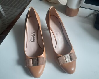 May include: A pair of tan leather Salvatore Ferragamo pumps. Each shoe features a bow detail with a metal buckle and the brand name embossed on the insole. The heels are approximately 2 inches high.