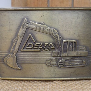 May include: A brass belt buckle featuring a Case Delta Series excavator. The buckle is rectangular and has a textured surface. The excavator is depicted in a raised design with the text "CASE" and "DELTA SERIES" visible.