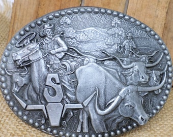 May include: Silver-toned metal belt buckle featuring a cowboy riding a horse and a herd of cattle. The buckle has a raised design and a textured finish.