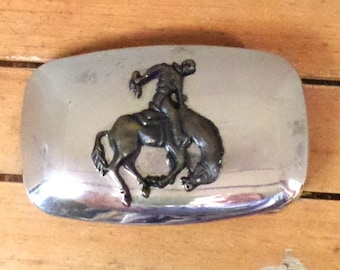 May include: Silver belt buckle with a raised design of a cowboy riding a bucking bronco.