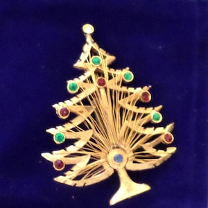 May include: A gold-toned Christmas tree brooch with red, green, and blue jewel accents. The brooch has a detailed design with a fan-like structure and a tree trunk base. The background is a dark blue velvet.