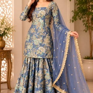 Op de afbeelding: Een blauwe tuniek met lange mouwen en een bijpassende rok tot op de grond met een ingewikkeld bloemen- en paisleypatroon in goud-, groenblauw- en wittinten. De outfit bevat een doorschijnende blauwe dupatta met een geborduurde gouden rand.