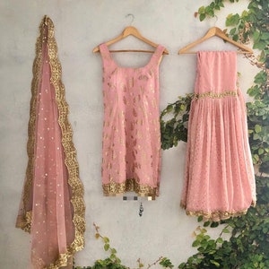 Peut inclure: Un ensemble trois pi&egrave;ces compos&eacute; d'une tunique rose sans manches avec des motifs floraux dor&eacute;s, une jupe pliss&eacute;e assortie avec une bordure dor&eacute;e, et un dupatta rose transparent avec des sequins dor&eacute;s et un bord festonn&eacute; dor&eacute;.