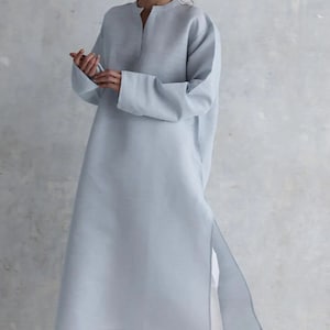 May include: A light blue long-sleeved tunic featuring a mandarin collar, a split neckline, and side slits, paired with white trousers and brown slide sandals. The tunic has a straight silhouette and reaches below the knees, made of a solid fabric.