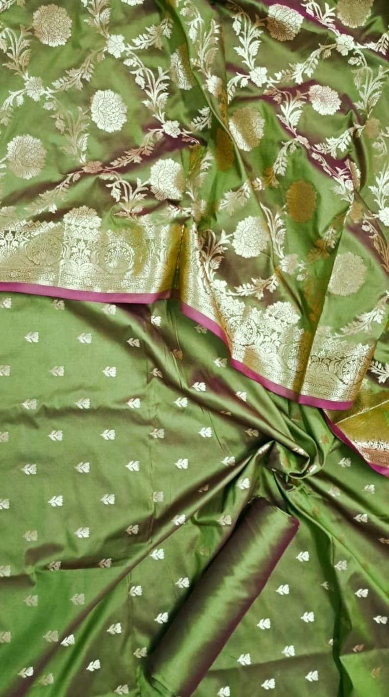 May include: Green silk fabric with a floral pattern. The fabric has a silver metallic thread woven into the design. The fabric is folded in half and the edges are visible.