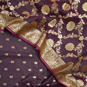 May include: A purple silk saree with a gold floral pattern and a maroon border. The saree is folded and draped over a matching purple fabric with a smaller gold floral pattern.