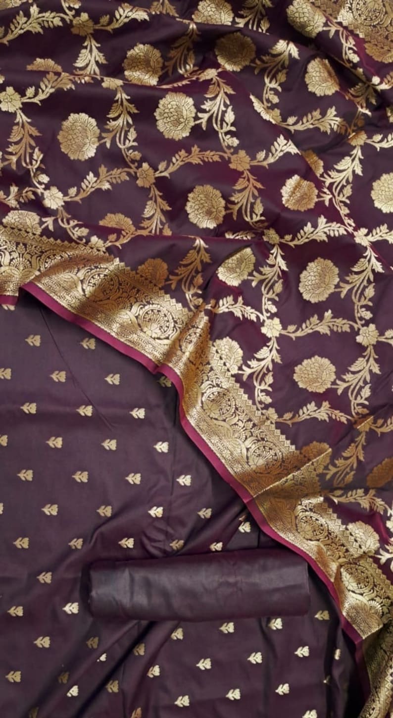 May include: A purple silk saree with a gold floral pattern and a maroon border. The saree is folded and draped over a matching purple fabric with a smaller gold floral pattern.