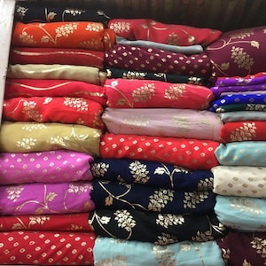 Peut inclure: Une collection de rouleaux de tissu pli&eacute;s empil&eacute;s en rang&eacute;es, pr&eacute;sentant une vari&eacute;t&eacute; de couleurs vives dont le rouge, l'orange, le rose, le bleu et le noir. Chaque pi&egrave;ce de textile est orn&eacute;e de motifs floraux ou g&eacute;om&eacute;triques dor&eacute;s.