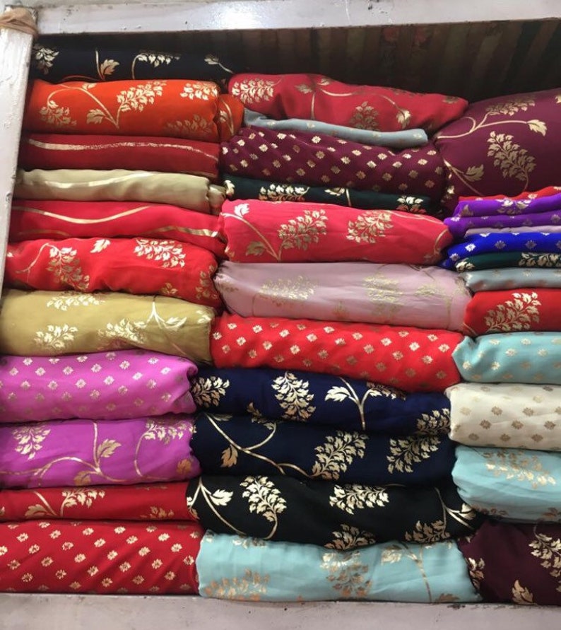 Peut inclure: Une collection de rouleaux de tissu pli&eacute;s empil&eacute;s en rang&eacute;es, pr&eacute;sentant une vari&eacute;t&eacute; de couleurs vives dont le rouge, l'orange, le rose, le bleu et le noir. Chaque pi&egrave;ce de textile est orn&eacute;e de motifs floraux ou g&eacute;om&eacute;triques dor&eacute;s.