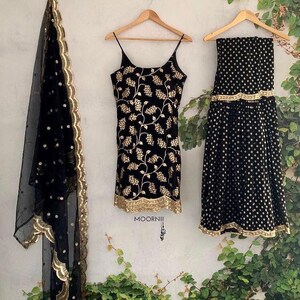 Peut inclure: Un ensemble trois pi&egrave;ces noir comprenant un haut sans manches avec broderies florales dor&eacute;es, une jupe assortie &agrave; ourlet dor&eacute; et un dupatta noir transparent &agrave; pois dor&eacute;s avec une bordure dor&eacute;e festonn&eacute;e. Le texte MOORNII est visible.