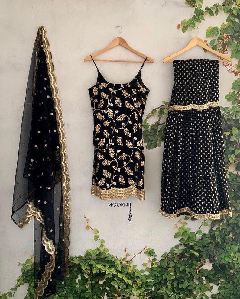Peut inclure: Un ensemble trois pi&egrave;ces noir comprenant un haut sans manches avec broderies florales dor&eacute;es, une jupe assortie &agrave; ourlet dor&eacute; et un dupatta noir transparent &agrave; pois dor&eacute;s avec une bordure dor&eacute;e festonn&eacute;e. Le texte MOORNII est visible.
