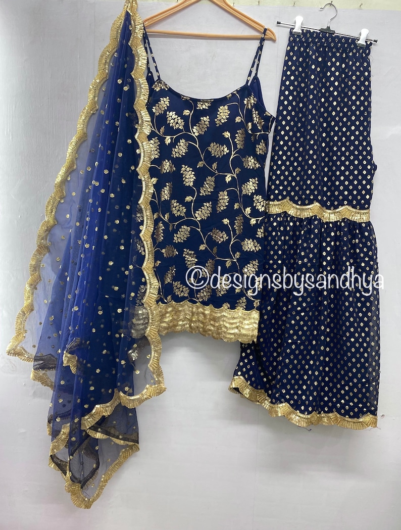 Peut inclure: Un haut sans manches bleu marine &agrave; motifs floraux dor&eacute;s, un pantalon large assorti &agrave; imprim&eacute; g&eacute;om&eacute;trique dor&eacute; et une dupatta bleu marine transparente avec une bordure festonn&eacute;e dor&eacute;e. Le texte designsbysandhya est visible.
