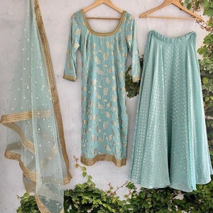 Peut inclure: Un ensemble trois pi&egrave;ces bleu clair compos&eacute; d'une tunique &agrave; manches longues avec une encolure d&eacute;gag&eacute;e, d'une jupe &eacute;vas&eacute;e et d'un dupatta transparent. La tunique et la jupe pr&eacute;sentent un motif floral dor&eacute;, tandis que le dupatta a une bordure dor&eacute;e.