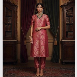 May include: A knee-length pink tunic with a gold floral brocade pattern, featuring short sleeves and a round neckline, paired with matching pink straight-leg trousers. The outfit is accessorized with a gold necklace and gold bangles.