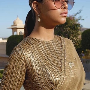 Könnte beinhalten: Eine Frau trägt eine goldene Paillettenbluse mit einem braunen, durchsichtigen Sari. Die Bluse hat einen runden Ausschnitt und einen gewellten Saum. Die Frau trägt eine Sonnenbrille und Ohrringe.