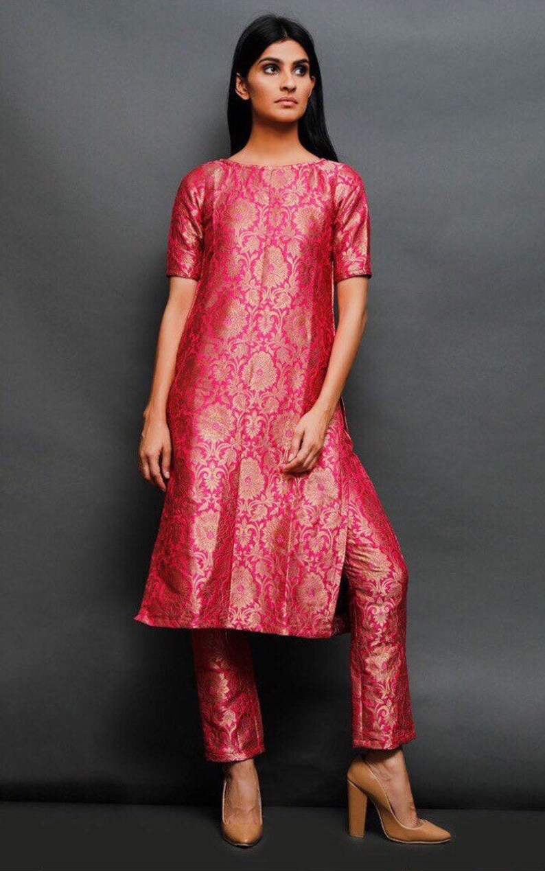 May include: A bright pink and gold brocade tunic with short sleeves and a round neckline, paired with matching straight-leg trousers. The fabric features an intricate floral pattern woven throughout, creating a cohesive two-piece set.
