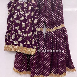 Peut inclure: Un ensemble haut sans manches et pantalon large violet fonc&eacute; avec une broderie florale dor&eacute;e sur le haut et un motif g&eacute;om&eacute;trique dor&eacute; sur le pantalon. Les deux pi&egrave;ces ont une bordure dor&eacute;e festonn&eacute;e aux ourlets. Le texte designsbysandhya est visible.
