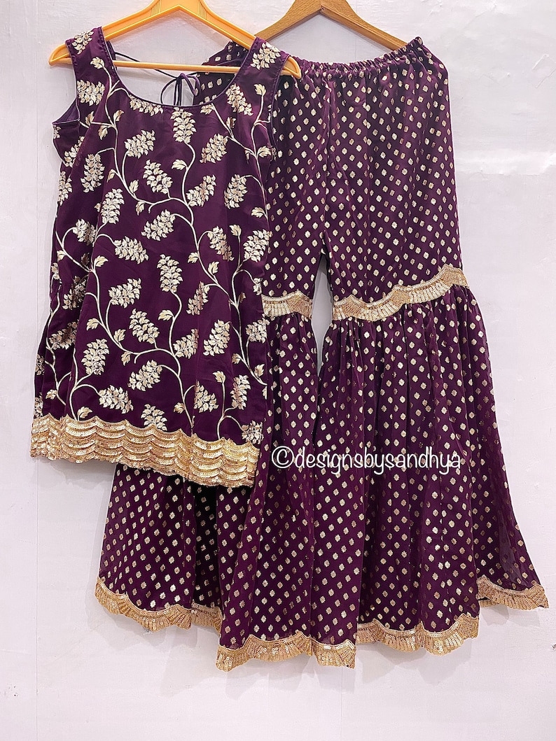 Peut inclure: Un ensemble haut sans manches et pantalon large violet fonc&eacute; avec une broderie florale dor&eacute;e sur le haut et un motif g&eacute;om&eacute;trique dor&eacute; sur le pantalon. Les deux pi&egrave;ces ont une bordure dor&eacute;e festonn&eacute;e aux ourlets. Le texte designsbysandhya est visible.