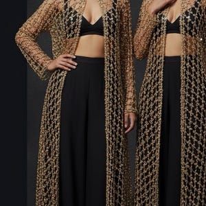 Puede incluir: Dos modelos con conjuntos negros a juego con chaquetas largas doradas de tejido abierto. Las chaquetas tienen un delicado patrón de red. Cada modelo también lleva un collarín y una pieza para la cabeza dorados. El fondo es gris oscuro.