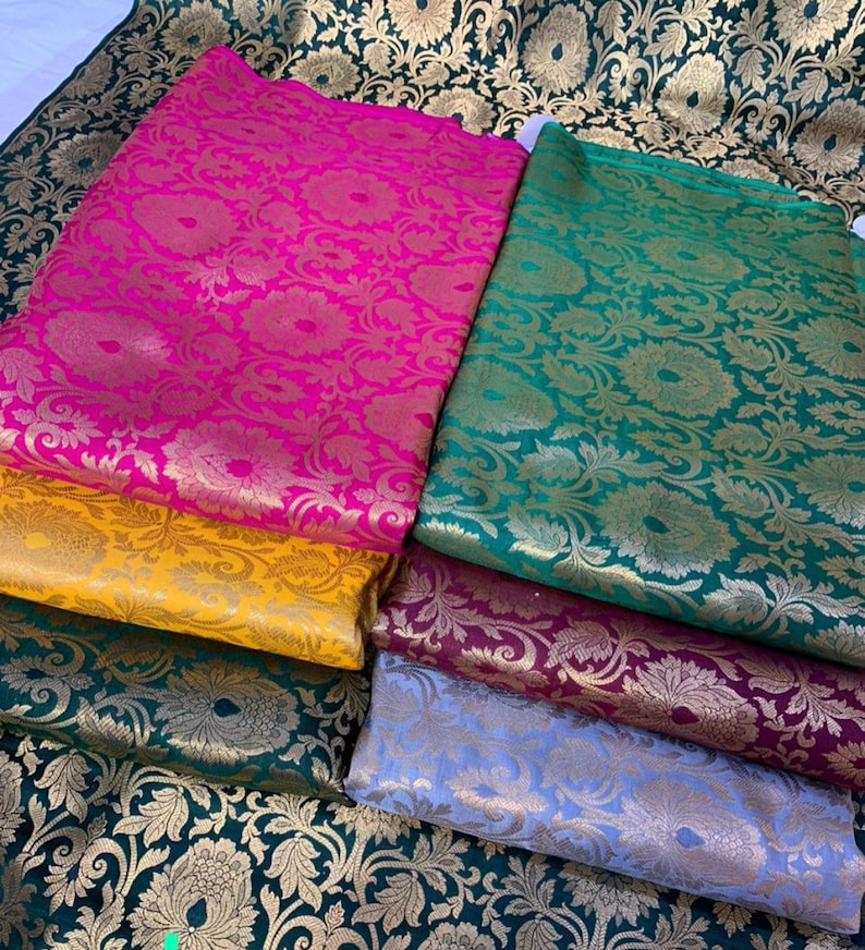 May include: A collection of folded fabric pieces featuring a repeating floral and leaf pattern in metallic gold thread. The fabrics are available in vibrant colors including hot pink, mustard yellow, dark green, deep maroon, and light lavender.