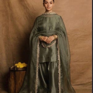 May include: A sage green salwar kameez set featuring a long-sleeved tunic, wide-leg trousers, and a sheer dupatta. The ensemble is crafted from a smooth, lustrous fabric and is accented with intricate, metallic-toned beaded trim along the edges.