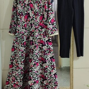 Pu&ograve; includere: Un abito nero a maniche lunghe con un vivace motivo floreale rosa e bianco, abbinato a pantaloni neri. L'abito presenta un dettagliato design a paisley e un bordo dorato.