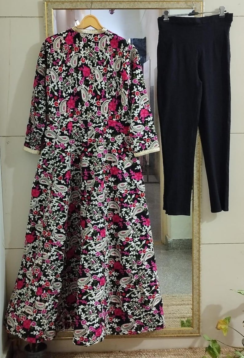 Pu&ograve; includere: Un abito nero a maniche lunghe con un vivace motivo floreale rosa e bianco, abbinato a pantaloni neri. L'abito presenta un dettagliato design a paisley e un bordo dorato.