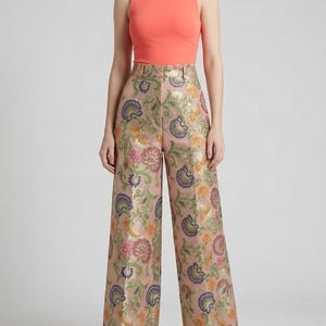 May include: A sleeveless coral-colored top paired with high-waisted, wide-leg trousers. The trousers feature a light pink base with an intricate, multi-colored floral and paisley jacquard pattern in shades of orange, purple, and metallic gold.