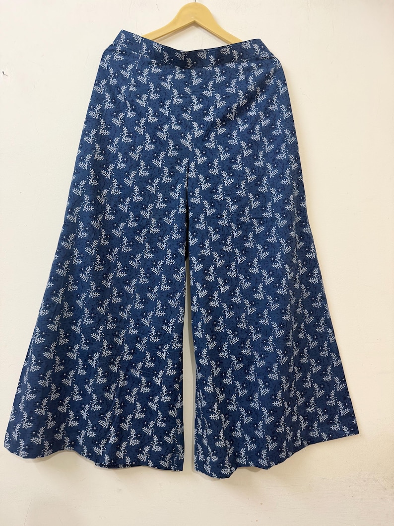 May include: Wide-leg trousers in a deep blue color featuring a repeating white botanical pattern of small leaves and tiny flowers. The pants have a high waistband and a loose, flowing silhouette that flares out from the hips to the hemline.