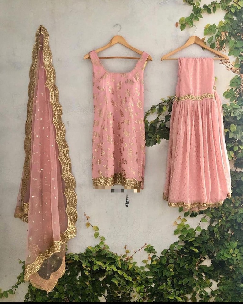 Peut inclure: Un ensemble trois pi&egrave;ces compos&eacute; d'une tunique rose sans manches avec des motifs floraux dor&eacute;s, une jupe pliss&eacute;e assortie avec une bordure dor&eacute;e, et un dupatta rose transparent avec des sequins dor&eacute;s et un bord festonn&eacute; dor&eacute;.