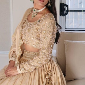 Könnte beinhalten: Ein goldfarbenes, verziertes formelles Kleid mit langen Ärmeln. Das Oberteil und die Ärmel sind mit aufwendigen Perlenarbeiten verziert. Das Outfit beinhaltet einen passenden Rock und einen zarten Schal. Das Ensemble wird durch ein weißes Halsband und grüne Ohrringe ergänzt.