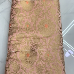 May include: A folded piece of fabric in a soft pink hue featuring an intricate woven pattern of gold floral motifs and leaves. Small, vibrant orange accents are interspersed throughout the design, creating a detailed and textured appearance.