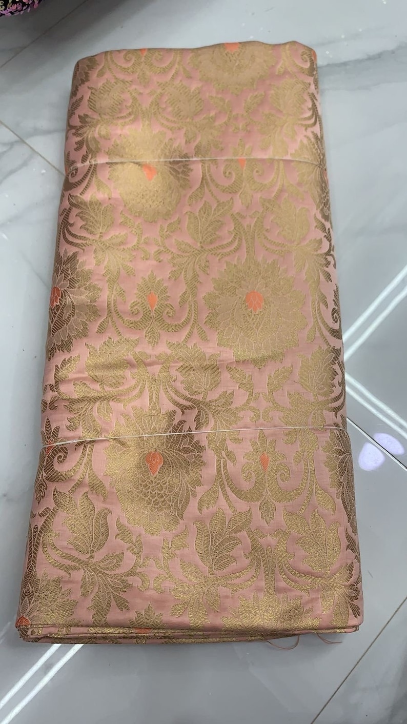 May include: A folded piece of fabric in a soft pink hue featuring an intricate woven pattern of gold floral motifs and leaves. Small, vibrant orange accents are interspersed throughout the design, creating a detailed and textured appearance.