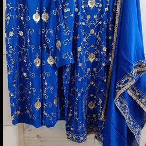 May include: A royal blue embroidered outfit, including a tunic, trousers, and a scarf. The tunic and trousers are adorned with gold floral embroidery. The scarf has gold trim. The outfit is displayed on a wooden hanger.