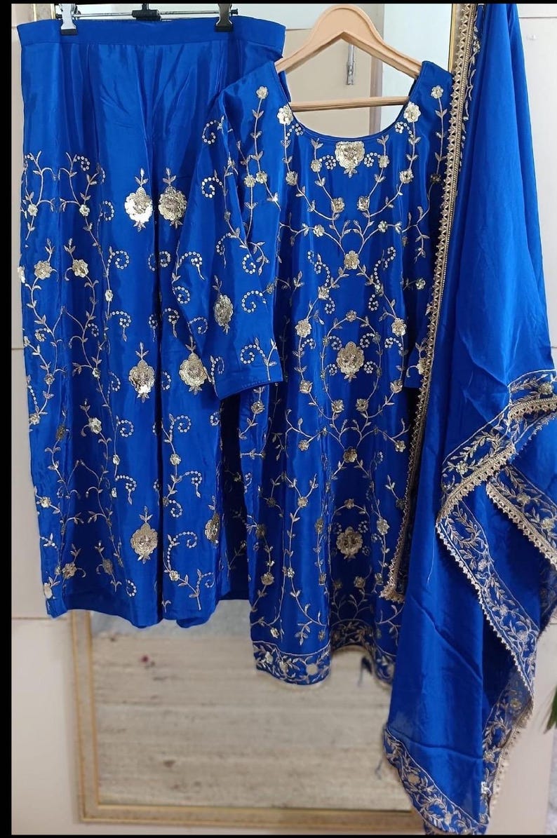 May include: A royal blue embroidered outfit, including a tunic, trousers, and a scarf. The tunic and trousers are adorned with gold floral embroidery. The scarf has gold trim. The outfit is displayed on a wooden hanger.